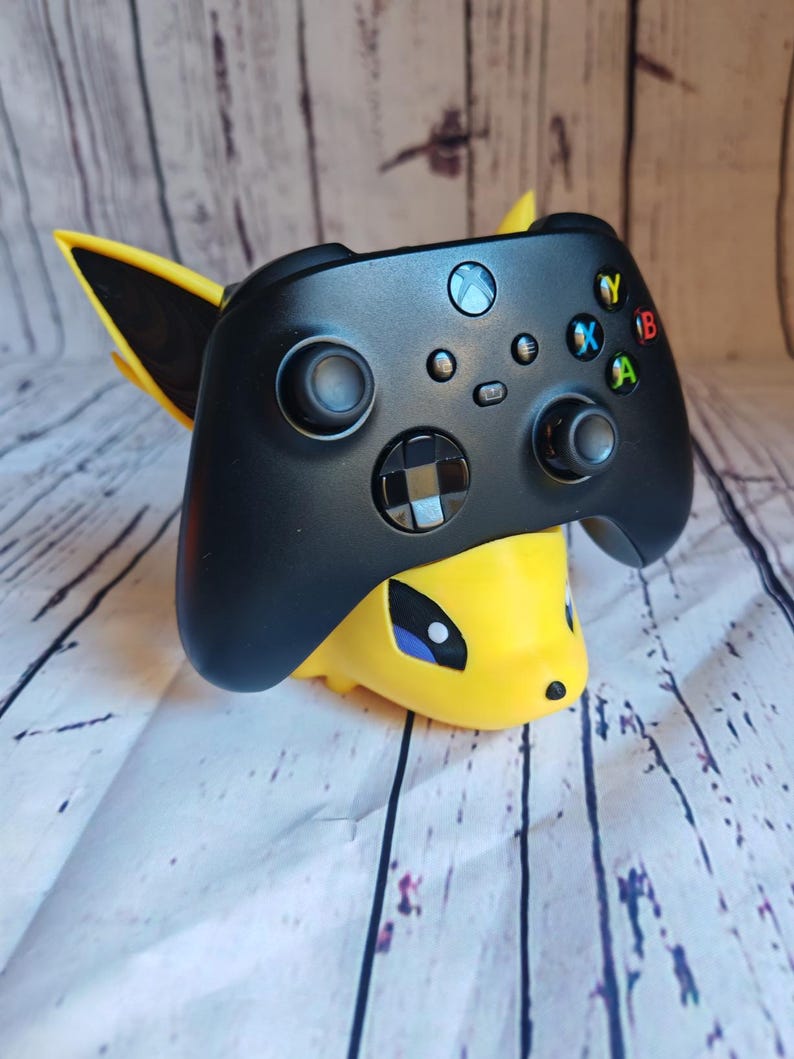 Peut inclure: Une manette Xbox noire est plac&eacute;e dans un support imprim&eacute; en 3D jaune et noir en forme de personnage de dessin anim&eacute;. Le support a un &oelig;il bleu et un nez jaune.