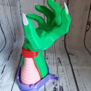 Op de afbeelding: Een groene hand met lange witte klauwen, die omhoog reikt. De hand is bevestigd aan een roze en paarse basis.