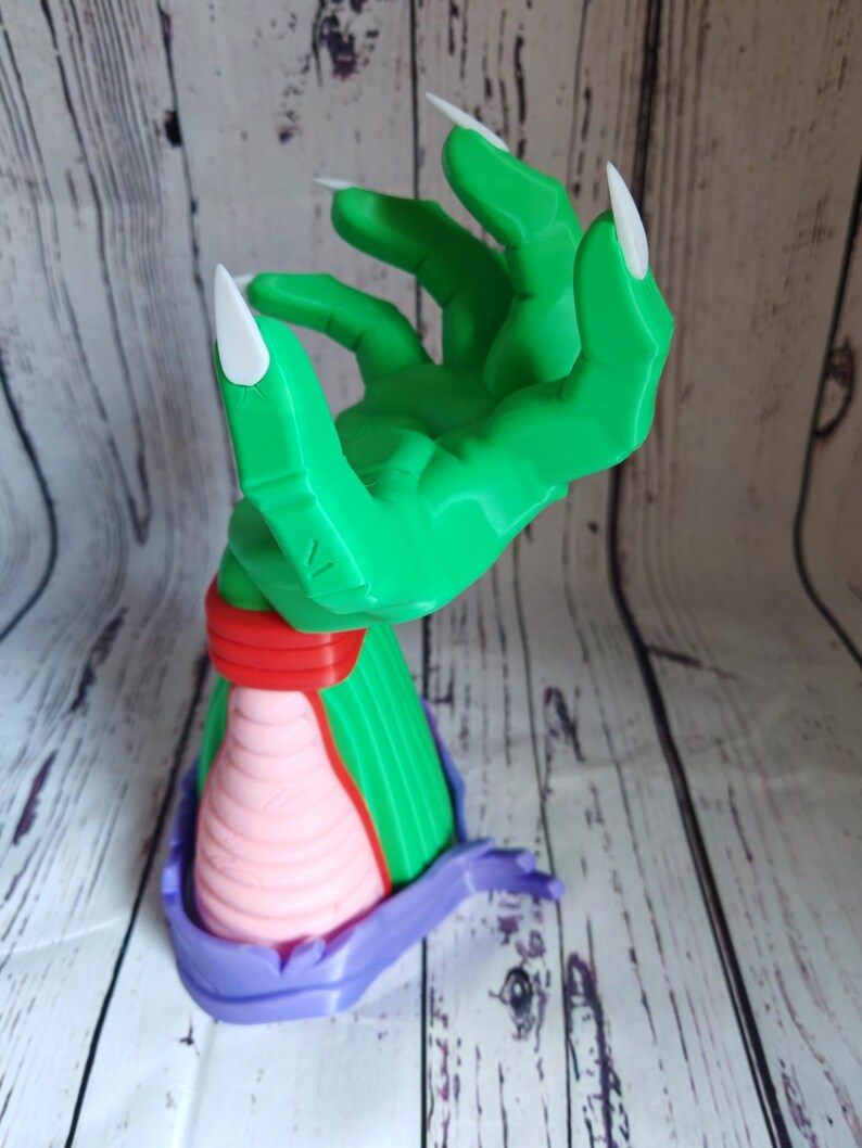 Op de afbeelding: Een groene hand met lange witte klauwen, die omhoog reikt. De hand is bevestigd aan een roze en paarse basis.