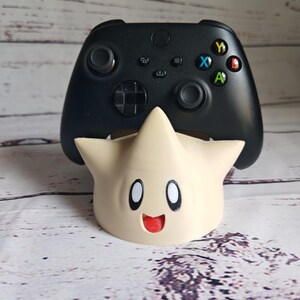 May include: A black video game controller is sitting on a beige 3D printed stand shaped like a star with a smiling face.