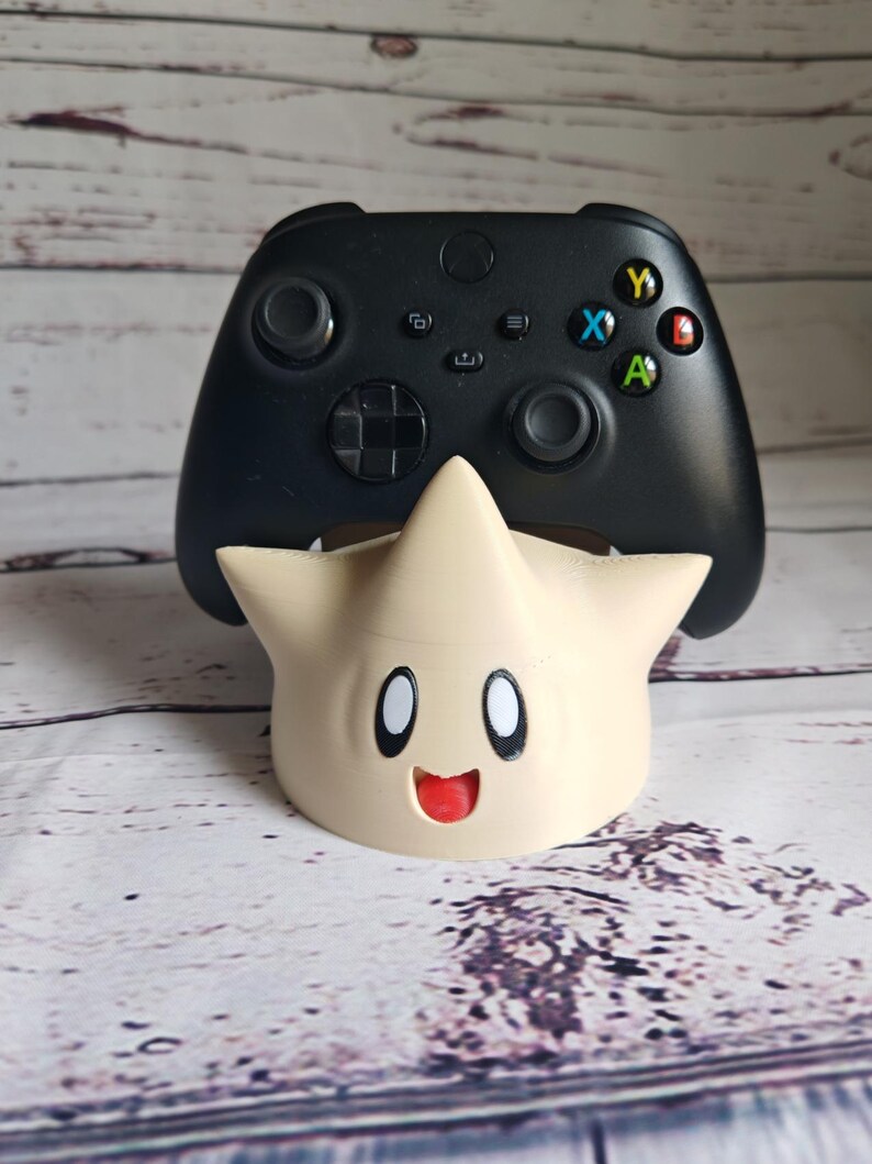 May include: A black video game controller is sitting on a beige 3D printed stand shaped like a star with a smiling face.