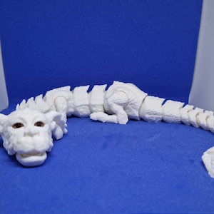 Op de afbeelding: Een wit, gearticuleerd drakenbeeldje met bruine ogen. De draak ligt op zijn buik met zijn kop naar de zijkant gedraaid. Het beeldje is gemaakt van plastic en heeft een gesegmenteerd lichaam.