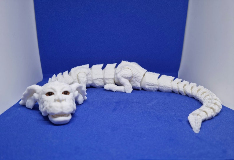 Op de afbeelding: Een wit, gearticuleerd drakenbeeldje met bruine ogen. De draak ligt op zijn buik met zijn kop naar de zijkant gedraaid. Het beeldje is gemaakt van plastic en heeft een gesegmenteerd lichaam.
