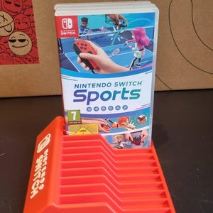 May include: A red plastic stand for Nintendo Switch game cartridges. The stand has the words "Nintendo Switch" printed on the side. A Nintendo Switch game cartridge for "Nintendo Switch Sports" is in the stand.