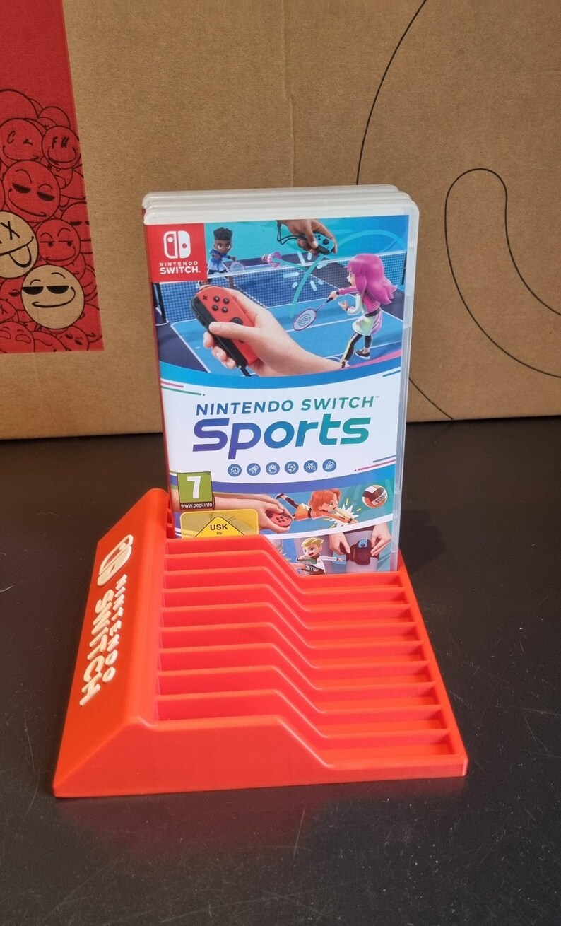 May include: A red plastic stand for Nintendo Switch game cartridges. The stand has the words "Nintendo Switch" printed on the side. A Nintendo Switch game cartridge for "Nintendo Switch Sports" is in the stand.