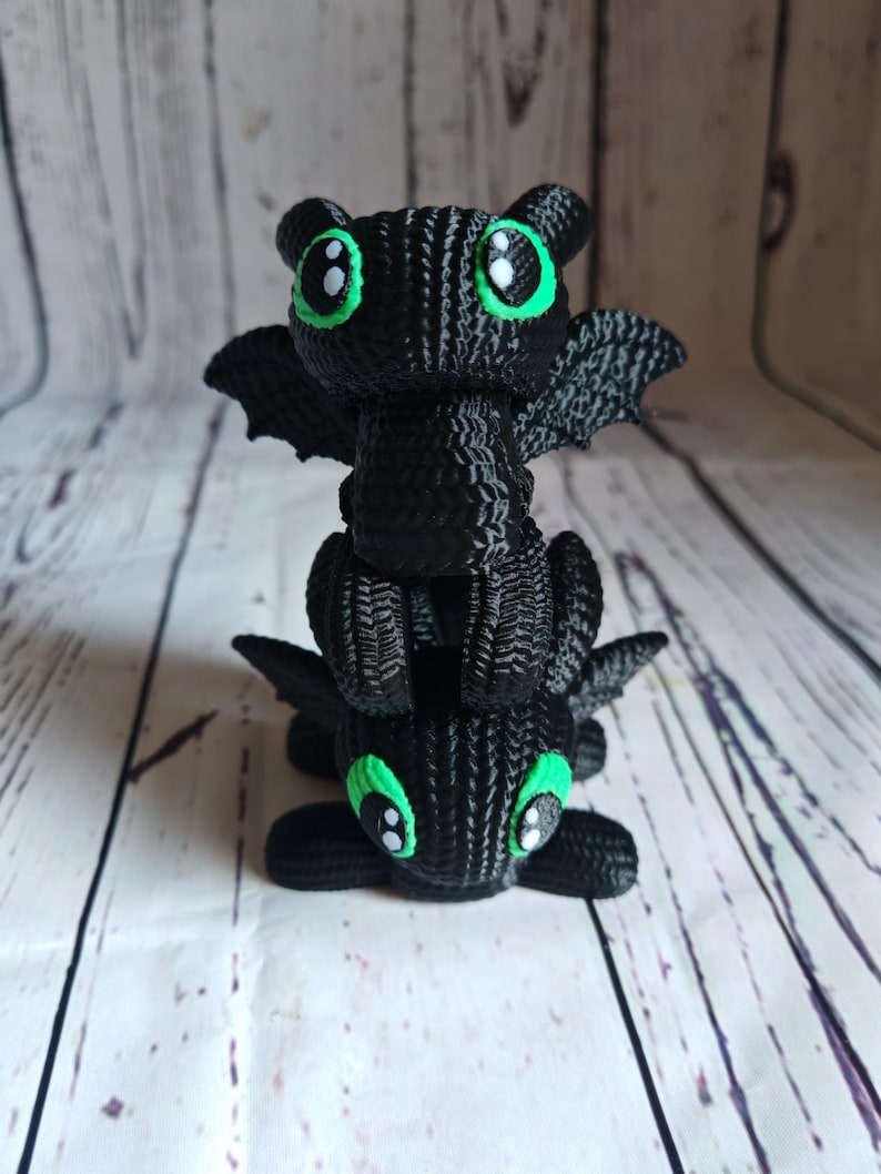 May include: A stack of two black knitted dragon figurines with green eyes. The dragons are facing forward with their wings spread out.