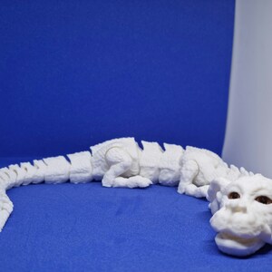 Op de afbeelding: Wit 3D-geprint model van een draak met een gesegmenteerd lichaam en een grote kop. De draak ligt op een blauwe ondergrond.