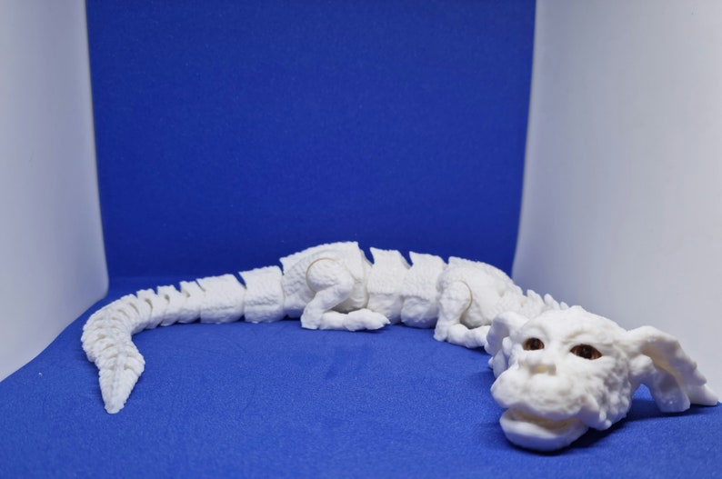 Op de afbeelding: Wit 3D-geprint model van een draak met een gesegmenteerd lichaam en een grote kop. De draak ligt op een blauwe ondergrond.