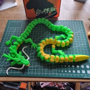 Op de afbeelding: Een groen en geel gearticuleerd draken speelgoed. De draak is gemaakt van plastic en heeft een lange staart, een kop met een witte tong en een reeks verbonden segmenten die het mogelijk maken om hem in verschillende poses te plaatsen.