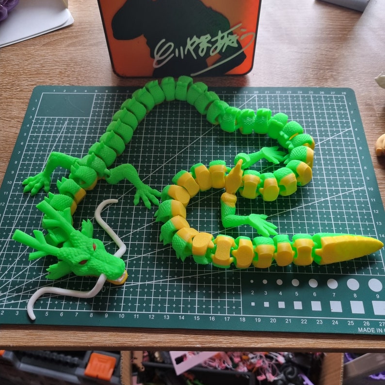 Op de afbeelding: Een groen en geel gearticuleerd draken speelgoed. De draak is gemaakt van plastic en heeft een lange staart, een kop met een witte tong en een reeks verbonden segmenten die het mogelijk maken om hem in verschillende poses te plaatsen.