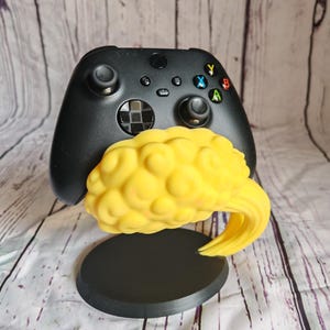 Op de afbeelding: Een zwarte Xbox-controller wordt getoond met een gele, hersenvormige standaard. De standaard heeft een zwarte cirkelvormige basis en een gebogen steun. De controller heeft gekleurde knoppen en joysticks.