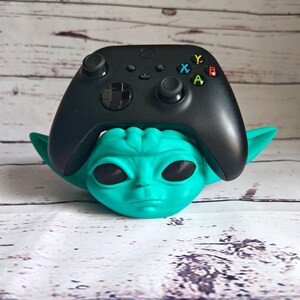 Op de afbeelding: Een tealgroen Yoda-achtig figuur dat een zwarte Xbox-controller vasthoudt. De controller heeft de standaard Xbox-knoppen: A, B, X, Y.