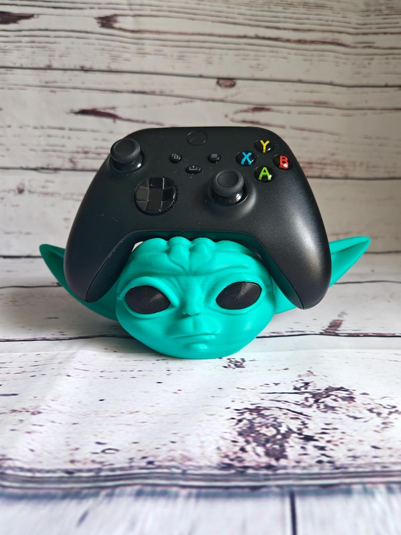 Op de afbeelding: Een tealgroen Yoda-achtig figuur dat een zwarte Xbox-controller vasthoudt. De controller heeft de standaard Xbox-knoppen: A, B, X, Y.