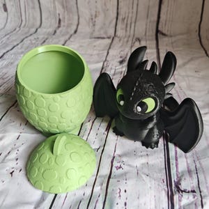 K&ouml;nnte beinhalten: Eine schwarze Drachenfigur mit gr&uuml;nen Augen und Fl&uuml;geln steht neben einem hellgr&uuml;nen, eif&ouml;rmigen Beh&auml;lter mit Deckel. Der Beh&auml;lter und der Deckel haben ein strukturiertes, gepunktetes Muster. Der Drache ist eine Figur aus der "Drachenz&auml;hmen leicht gemacht"-Reihe.