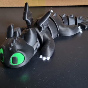 May include: A black 3D printed dragon figurine with green eyes and white claws. The dragon is lying on its side with its head resting on the ground.