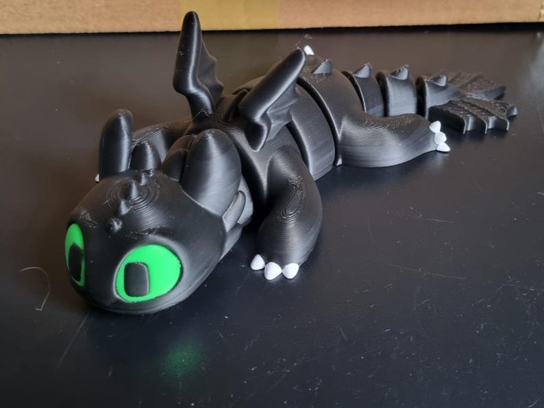 May include: A black 3D printed dragon figurine with green eyes and white claws. The dragon is lying on its side with its head resting on the ground.