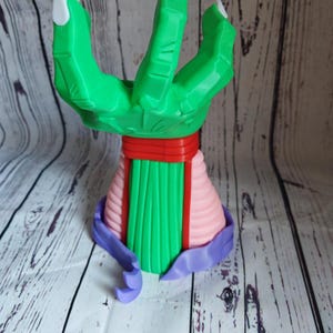 Op de afbeelding: Een groene hand met drie vingers, witte klauwen, een rode band en een roze en groene basis. De hand is gemaakt van plastic en is een 3D-geprint model.