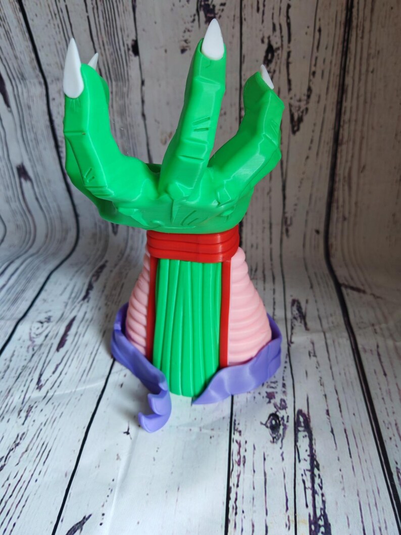 Op de afbeelding: Een groene hand met drie vingers, witte klauwen, een rode band en een roze en groene basis. De hand is gemaakt van plastic en is een 3D-geprint model.