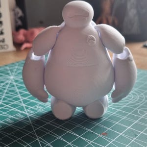 May include: White 3D printed figurine of Baymax, a large, round, white robot character with arms outstretched. The figurine is standing on a green cutting mat.