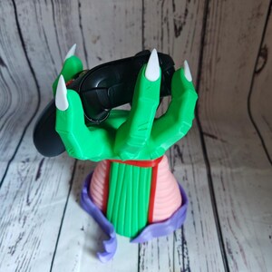 Op de afbeelding: Een groene hand met drie vingers en witte klauwen houdt een zwarte videogamecontroller vast. De hand is bevestigd aan een roze en groene basis.