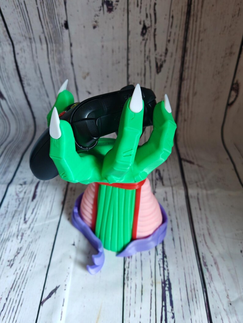 Op de afbeelding: Een groene hand met drie vingers en witte klauwen houdt een zwarte videogamecontroller vast. De hand is bevestigd aan een roze en groene basis.