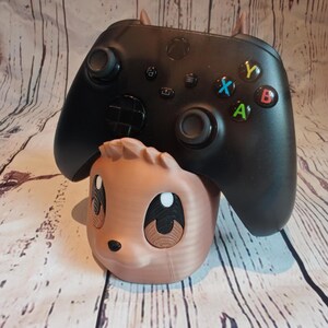 Peut inclure: Un support de manette imprim&eacute; en 3D de couleur marron et noir en forme d'animal de dessin anim&eacute; avec de grands yeux. Une manette Xbox noire est pos&eacute;e sur le support.