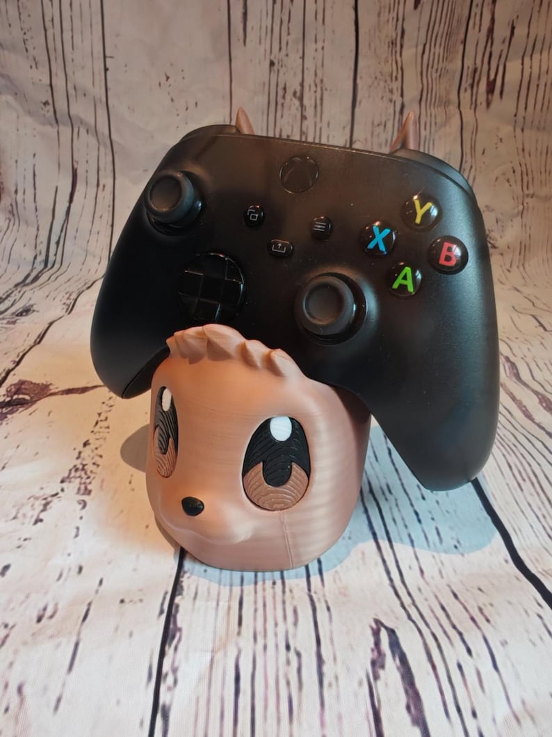 Peut inclure: Un support de manette imprim&eacute; en 3D de couleur marron et noir en forme d'animal de dessin anim&eacute; avec de grands yeux. Une manette Xbox noire est pos&eacute;e sur le support.