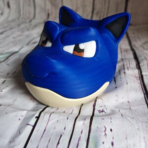 K&ouml;nnte beinhalten: Ein blaues, 3D-gedrucktes Blastoise-Kopf, mit wei&szlig;en Augen, braunen Pupillen, schwarzen Ohren und einem cremefarbenen Sockel. Die Skulptur steht vor einem Holzmaserungshintergrund.