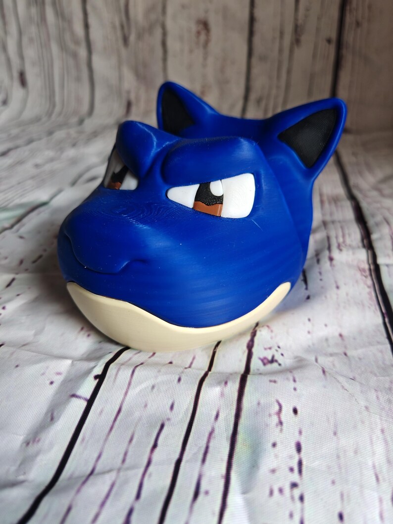 K&ouml;nnte beinhalten: Ein blaues, 3D-gedrucktes Blastoise-Kopf, mit wei&szlig;en Augen, braunen Pupillen, schwarzen Ohren und einem cremefarbenen Sockel. Die Skulptur steht vor einem Holzmaserungshintergrund.