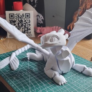 May include: A white 3D printed model of a dragon with wings outstretched. The dragon is posed in a sitting position with its head tilted up. The model is printed in a single color and has a smooth finish.