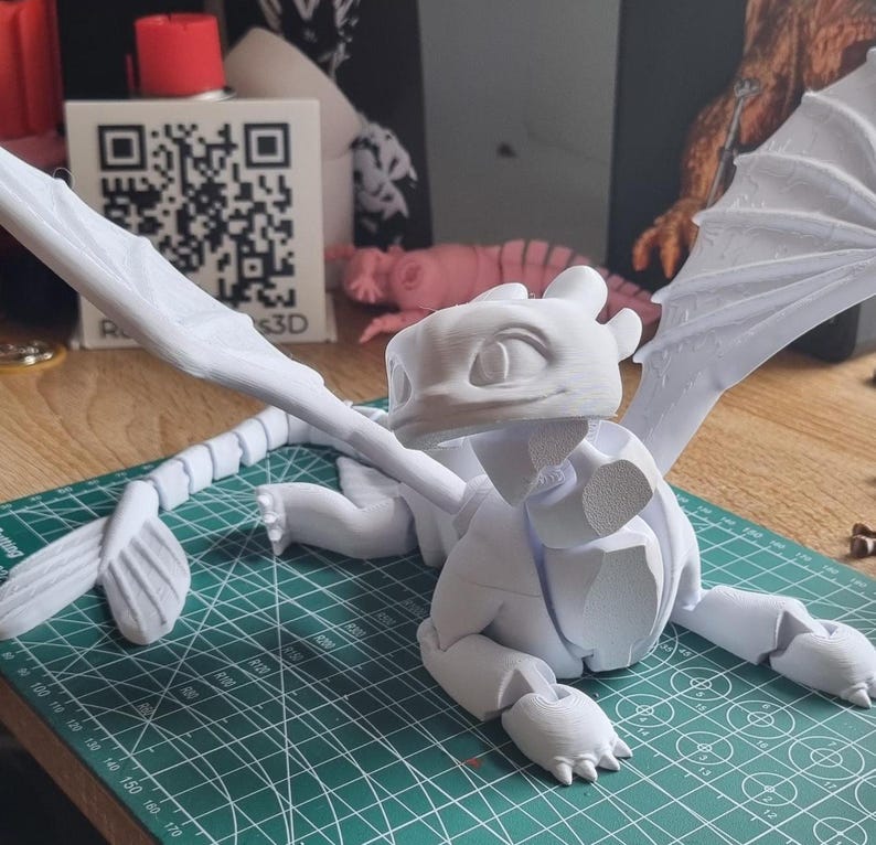 May include: A white 3D printed model of a dragon with wings outstretched. The dragon is posed in a sitting position with its head tilted up. The model is printed in a single color and has a smooth finish.