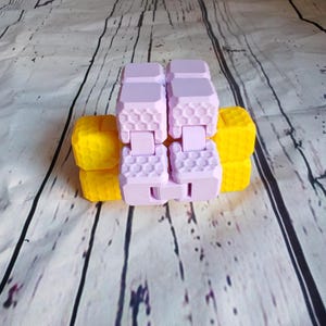 Peut inclure: Un jouet anti-stress rose et jaune avec un motif en nid d'abeille. Le jouet est en plastique et a une forme carr&eacute;e.