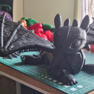 K&ouml;nnte beinhalten: Eine schwarze, 3D-gedruckte Drachenfigur mit gro&szlig;en Fl&uuml;geln, einem strukturierten K&ouml;rper und einem detaillierten Gesicht. Der Drache sitzt auf einer gr&uuml;nen Schneidematte.