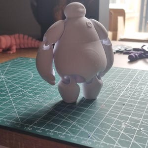May include: A white 3D printed figurine of Baymax, the inflatable healthcare companion from the Disney movie Big Hero 6. The figurine is standing on a green cutting mat.