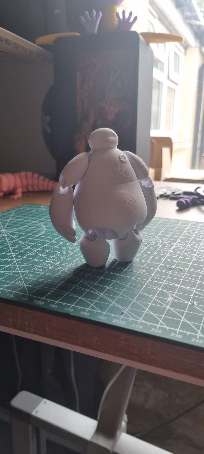 May include: A white 3D printed figurine of Baymax, the inflatable healthcare companion from the Disney movie Big Hero 6. The figurine is standing on a green cutting mat.