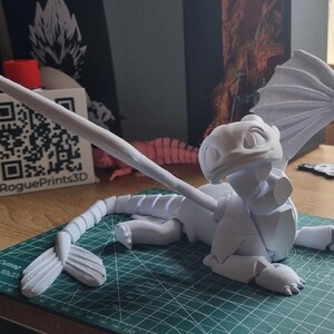 May include: A white 3D printed model of a dragon with wings, a tail, and a head with a friendly expression. The model is posed on a green cutting mat with a white background.