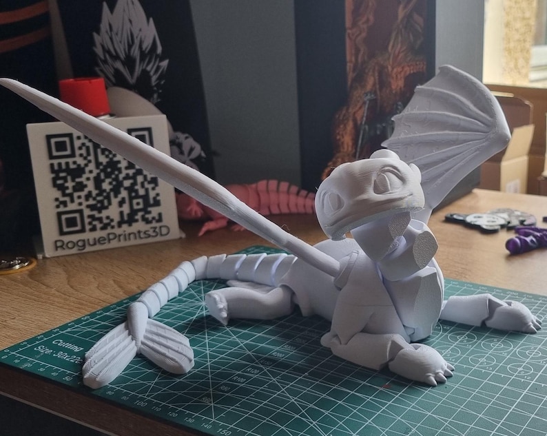 May include: A white 3D printed model of a dragon with wings, a tail, and a head with a friendly expression. The model is posed on a green cutting mat with a white background.