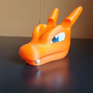 Op de afbeelding: Oranje plastic speelgoed drakenkop met een blauw oog. De draak heeft een tandenrij en is gemaakt van glad, mat plastic.