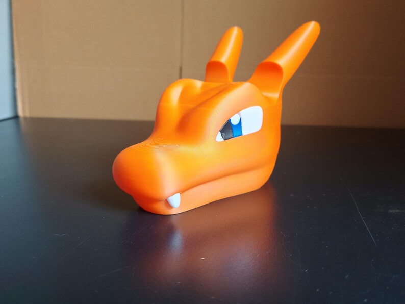 Op de afbeelding: Oranje plastic speelgoed drakenkop met een blauw oog. De draak heeft een tandenrij en is gemaakt van glad, mat plastic.