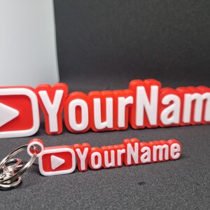 Puede incluir: Llavero impreso en 3D rojo y blanco con un bot&oacute;n de reproducci&oacute;n y el texto "Your Name".