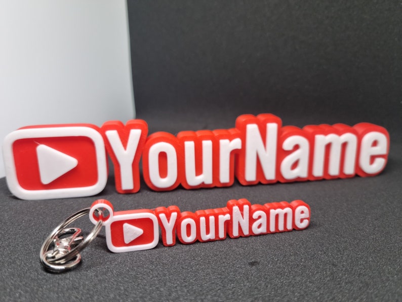 Puede incluir: Llavero impreso en 3D rojo y blanco con un bot&oacute;n de reproducci&oacute;n y el texto "Your Name".