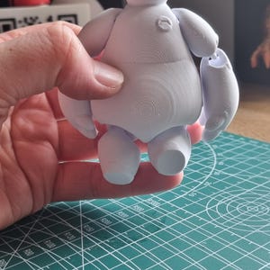 May include: A white 3D printed figurine of Baymax, the inflatable healthcare companion from the Disney movie Big Hero 6. The figurine is posed standing with its arms outstretched.