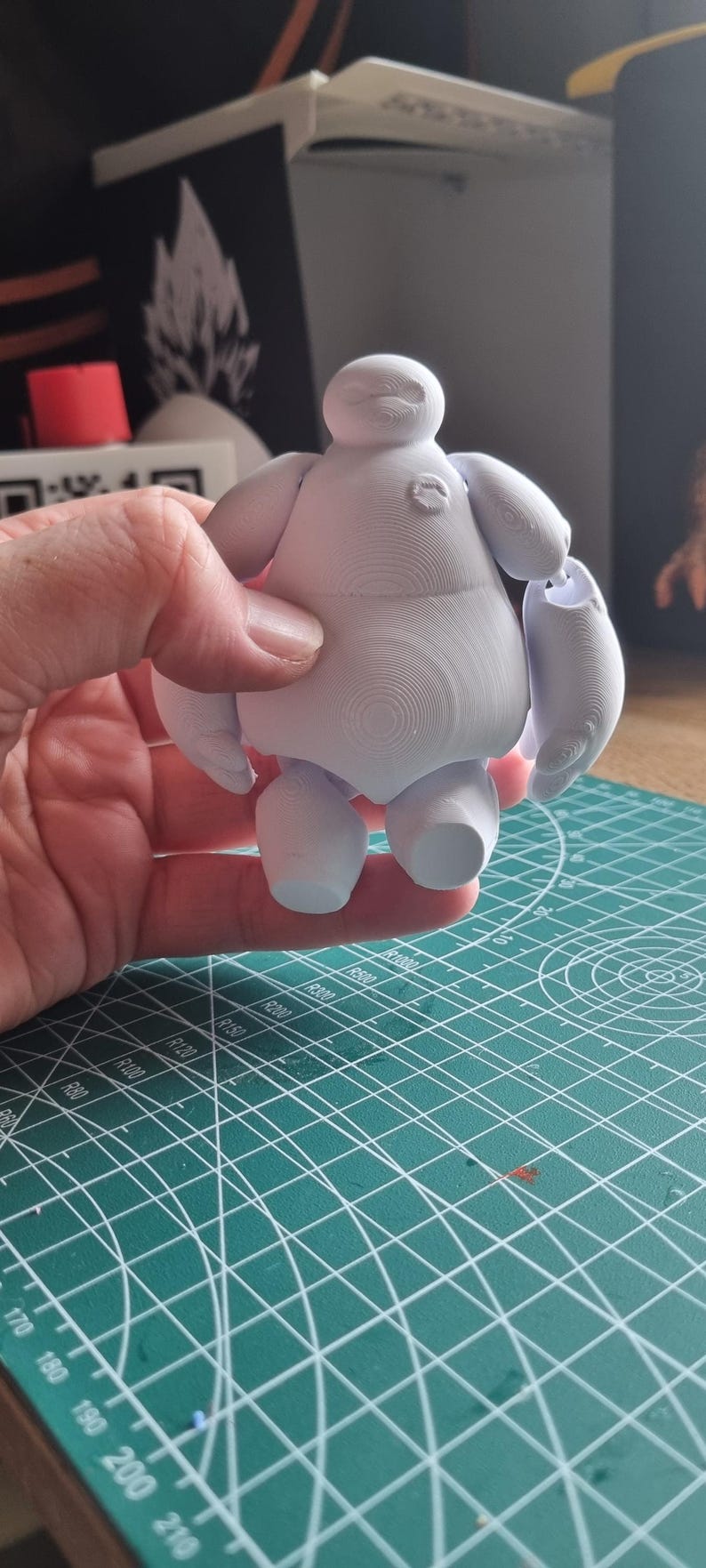May include: A white 3D printed figurine of Baymax, the inflatable healthcare companion from the Disney movie Big Hero 6. The figurine is posed standing with its arms outstretched.
