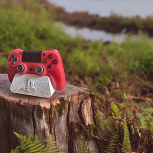 May include: A red video game controller sits on a gray and white stand with the PlayStation logo. The stand is on a tree stump in a forest setting.