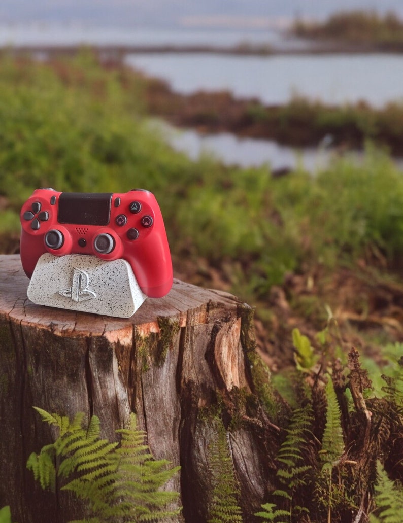 May include: A red video game controller sits on a gray and white stand with the PlayStation logo. The stand is on a tree stump in a forest setting.