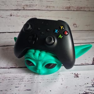 Op de afbeelding: Een groene en zwarte videogamecontrollerstandaard in de vorm van een personage uit de Star Wars-franchise. De standaard is gemaakt van plastic en heeft een getextureerd oppervlak. De controller is zwart en heeft een groene, gele, rode en blauwe knop.