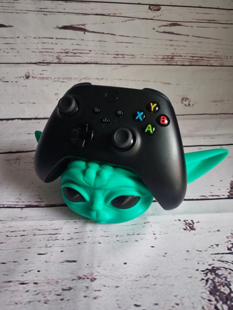 Op de afbeelding: Een groene en zwarte videogamecontrollerstandaard in de vorm van een personage uit de Star Wars-franchise. De standaard is gemaakt van plastic en heeft een getextureerd oppervlak. De controller is zwart en heeft een groene, gele, rode en blauwe knop.