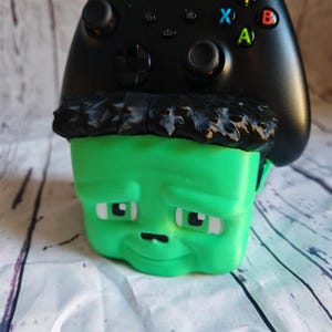 Könnte beinhalten: Ein grüner, 3D-gedruckter Controller-Halter in Form eines Cartoon-Gesichts mit schwarzen Haaren und einem schwarzen Xbox-Controller, der obenauf liegt. Der Controller hat farbige Tasten und eine matte Oberfläche.