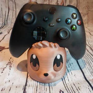 Peut inclure: Une figurine en 3D imprim&eacute;e de couleur marron et beige avec un visage de dessin anim&eacute; et de grands yeux. Une manette Xbox noire repose sur la figurine.