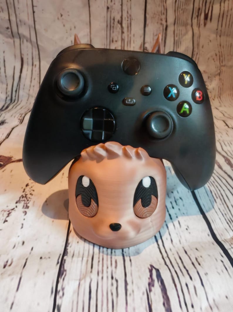 Peut inclure: Une figurine en 3D imprim&eacute;e de couleur marron et beige avec un visage de dessin anim&eacute; et de grands yeux. Une manette Xbox noire repose sur la figurine.